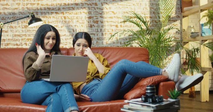 Two pretty Caucasian girls twins crying while watching drama movie on laptop on couch in room. Couple of beautiful sad young women wipe away tears while watching film on computer at sofa. Home concept