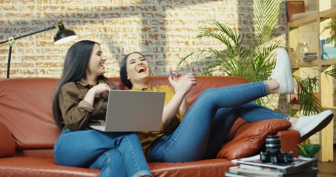 Couple of beautiful cheerful young women laughing while watching film on computer on red sofa. Two pretty Caucasian girls smiling while watching comedy movie on laptop on couch in room. Home concept