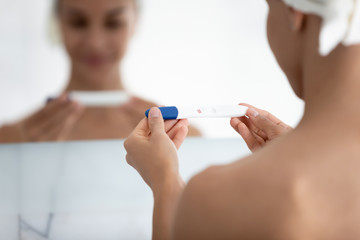 Woman reflected in mirror view over female shoulder, close up focus on hands holds pregnancy test with two red stripes result confirming gestation, rapid detection pregnancy tool advertisement concept