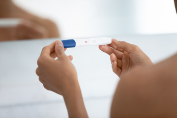 Close up view over shoulder of woman hold positive pregnancy quick plastic test with two red stripes confirm that female is pregnant. New life, feminine health, happy changes and fertilization concept
