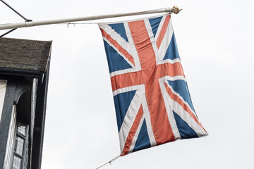 Obraz premium Flag of United Kingdom (the Union Jack also known as Union flag) blowing in the wind in front of an old traditional timber house, towards cloudy grey sky in a rainy day 