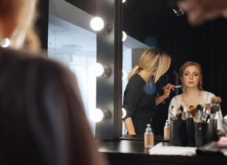Portrait of professional hairdresser with hairspray in hands working with client. hairdresser makes wedding hairstyle to beautiful blonde bride in front of mirror studio portrait. evening hairstyle
