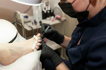 Pedicure in a beauty salon. A masked and gloved craftsman varnishes the client’s nails. Close-up.