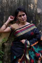 Portrait of a beautiful brunette Indian bengali thoughtful woman in traditional wear sari standing beside a tree on a drenched morning of Durga Puja festival in textured background. Indian lifestyle.