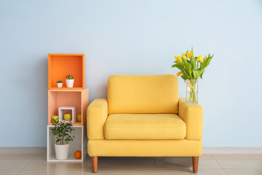 Interior Of Modern Room With Comfortable Armchair, Shelf Unit And Spring Flowers Near Light Wall