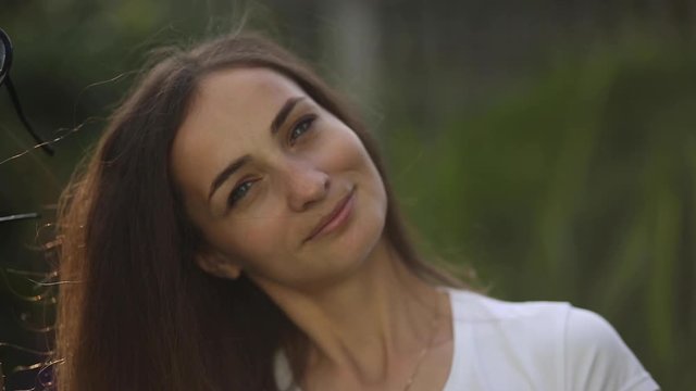 Close-up headshot stock video of a gorgeous pretty happy brunette Caucasian woman with thick hair smiling at camera. Unfocused blurred green trees in the background. Positivity, cheerfulness