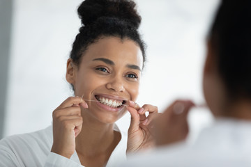 Beautiful smiling african ethnicity young woman in bathrobe looking at mirror holding dental floss...