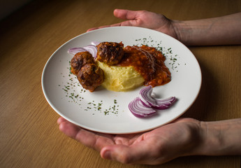 Woman hands holding plate with mashed potatoes and meatballs in tomato sauce on wooden table, side view