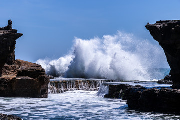Wave crashing onshore at low tide at Tanah Lot temple, Bali, Indonesia. Water cascading over stone, feeding tidal pool. 