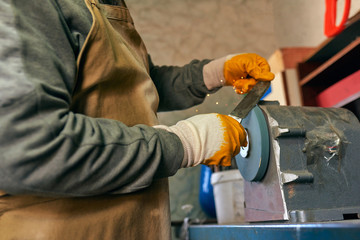 Fototapeta premium Horizontal side view image of adult man manufacturing in his workshop. Worker man sharpens the knife in the repair shop.
