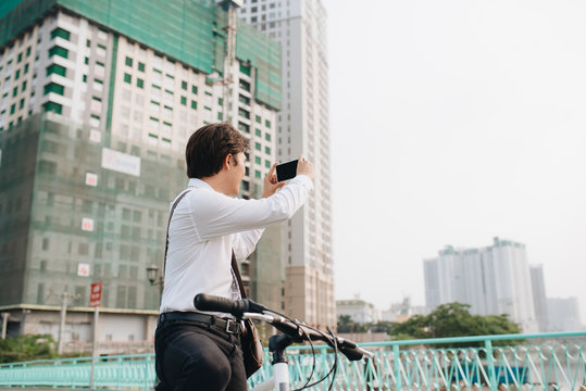 Smiling Asian man with bicycle taking photos in the city