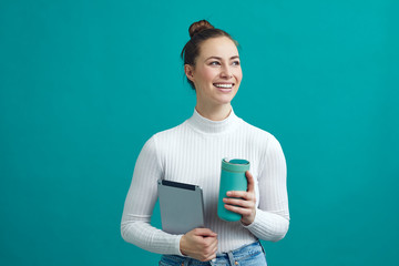 Attractive young woman standing on a colorful background, while holding a tablet and a thermocup...