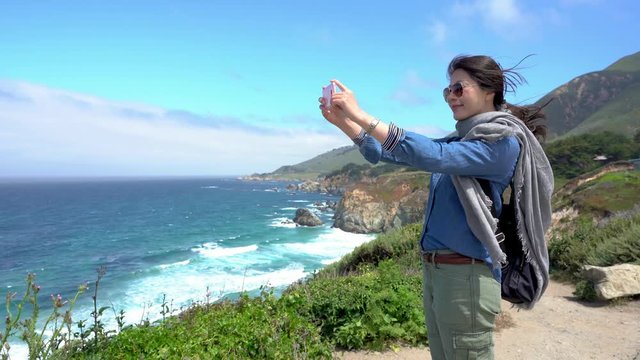 asian girl backpacker taking pictures of amazing landscape on mobile phone while standing on rock near sea. beautiful sunshine with sea and blue sky. Pacific coast highway 1 Big Sur California