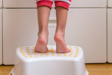 Small kids legs and feet tippy-toes on a step stool to reach for something on a cabinet counter.