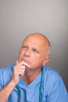 Doctor in pensive mood with hand on chin, wearing blue scrubs with stethoscope, against a light background.