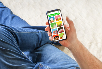Man in jeans lying on sofa and holding phone with app delivery food on screen. Food order concept.