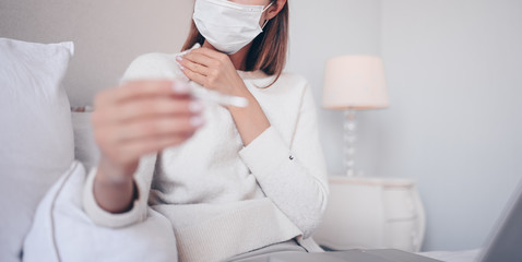 Fototapeta na wymiar Unrecognizable sick woman in face protection mask in bed holding thermometer at home quarantine isolation. Online work from home. Stay home safe concept. Suspecting Corona virus infection COVID-19. 