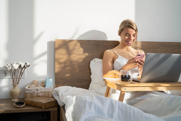 Beautiful freelancer girl working on a laptop and drinking coffee on a white bedsheet. Attractive blogger writes a blog article and has breakfast coffee and a croissant on brown wood table. top view