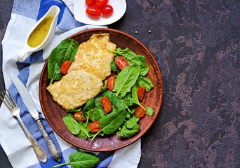Turkey breast chops on a brown clay plate on a dark concrete background. Served with a salad of fresh spinach and stewed cherry tomatoes. Top view, copy space.