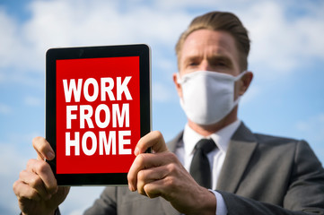 Businessman wearing coronavirus protective surgical face mask holding a tablet with a Work From Home message