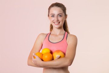 Young sports woman holding citrus fruits, lemon and orange in her hands, standing on a pink background and looking at the camera.