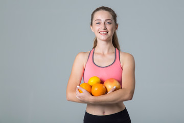 Young woman holds citrus fruits, lemon and orange in her hands, standing on a gray background and looking at the camera.