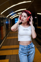 Young woman with burgundy pink maroon hair, in white top and light blue jeans, with red headphones, posing in subway, Three-quarter portrait of pretty girl in underground