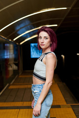 Young woman with burgundy red maroon short hair, wearing grey top and light blue jeans, posing in subway station, Three-quarter portrait of pretty girl at metro station