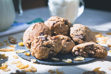 Plate with chocolate chip cookies, with glass of milk in the background on woody table with scattered cereals