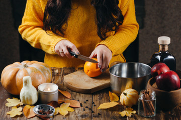 Close-up photo of woman in yellow sweater cutting orange for mulled wine. Autumn mood decorations on the table. Cozy athmosphere.