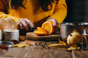 Close-up image of manicured woman's hands cutting oranges on wooden board for hot wine. Pumpkins, leaves, candles on the table. Autumn mod.