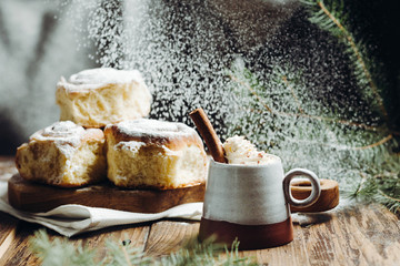 A hot cocoa with cinnamon stick and fresh Christmas buns with powder and over festive table, sugar powder is sprinkled on the background lke a snow. Winter holidays.