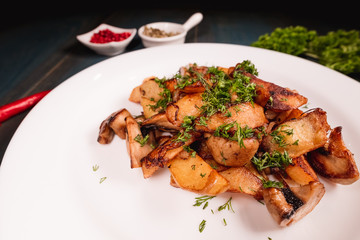 A plate of appetizing potato with mushrooms on a wooden table, among seasonings. Studio photography of food in the cooking industry, dark background.