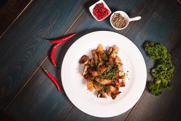 A plate of appetizing potato with mushrooms on a wooden table, among seasonings. Studio photography of food in the cooking industry, dark background.