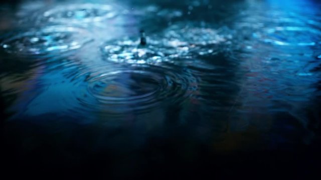 Rain drops falling into puddle on asphalt at night. Big city street lights mirror reflection on water surface. Close-up slow motion of raining outside in wet season