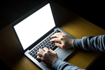 Man working on laptop with white screen glowing in the dark on a wooden desk from over the shoulder