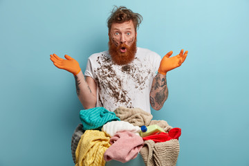 Puzzled dirty bearded ginger man housekeeper spreads palms and looks with stupor, poses near...
