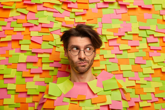 Photo of thoughtful European guy looks aside, has pensive expression, wears round spectacles, sticks out head from colorful background with adhesive notes, thinks about something interesting.