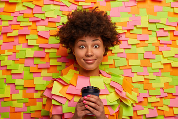 Glad dark skinned girl with Afro hair, holds paper cup of coffee, looks happily at camera, has surprised pleased expression, enjoys free time, sticks out head through background with adhesive notes