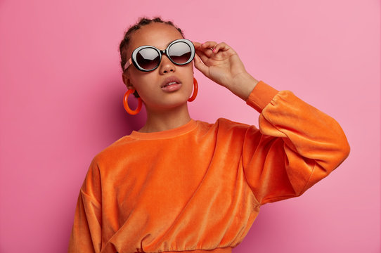 Photo of confident dark skinned Afro American female model wears trendy sunglasses, dressed in orange sweater, prepares for date, isolated on pink background, has fashion look, cornrow hairstyle