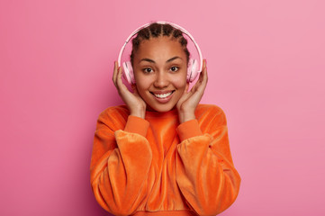 Happy teenage girl enjoys favourite music from playlist, wears stereo headphones, smiles gently, dressed in orange velvet sweater, feels energetic, isolated on pink background. People, lifestyle