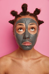Vertical photo of sad embarrassed woman concentrated above, wears facial clay mask for reducing acnes, stands naked, cares about body, isolated on pink background, has many combed buns on head