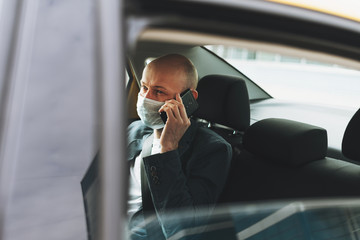 Bald man businessman in medical face mask using mobile phone inside yellow car taxi, concept of...
