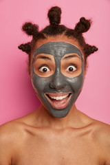 Pleased dark skinned woman has flawless skin, stares happily at camera, applies clay mask, stands nude against pink background, enjoys beauty treatments at home. Skin care, cosmetology concept