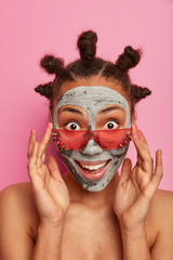 Positive young woman applies homemade mask on face, pampers skin, looks surprisingly through pink sunglasses, has naked body, bun hairstyle, stays beautiful, poses indoor. Anti age care concept