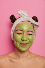 Skin care and beauty concept. Pleased mixed race young woman applies green mask for cleansing on face, cares about body, removes wrinkles, wears headband, stands topless against pink background