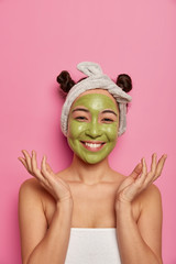 Happy Asian woman enjoys skin care routine and spa day at home, spreads palms and smiles gently, dressed in bath towel, has applied green face mask, stands against pink wall. Anti aging procedure