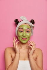 Beautiful pleased Asian woman cleans skin of face with green mask, wears headband and towel around body, enjoys cosmetic proedure in spa salon, models against pink background, free space above