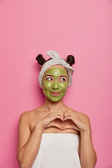 Facial treatment, cosmetology concept. Young satisfied Asian woman makes heart gesture, expresses affection, applies facial mask, wears headband and wrapped in towel, has fresh healthy skin.
