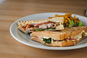 Close-up of chicken and bacon club sandwich with fries and vegetables on a white plate on a wooden background.